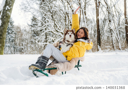 Young woman in yellow winter jacket sitting on a sled with a dog in snowy forest, surrounded by trees and snow-covered ground during a sunny winter day 140031368
