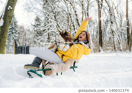 Young woman in yellow winter jacket joyfully sledding with a brown and white dog in a snowy forest, surrounded by trees and a winter landscape with bright sunlight 140031374