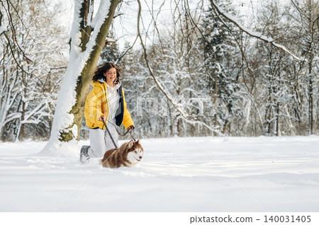 Woman in yellow winter jacket walking a dog on a leash through a snowy landscape with trees and a serene winter atmosphere during a sunny day 140031405