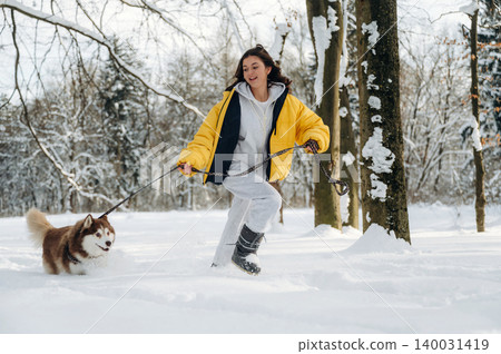 Young woman in a bright yellow jacket joyfully running with a brown and white dog on a snowy path surrounded by trees during winter outdoor activity Young woman in a bright yellow jacket joyfully running with a brown and white dog on a snowy path surrounded by trees during winter outdoor activity 140031419