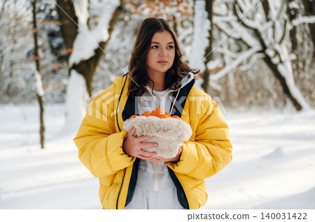 Young woman with long dark hair wearing a yellow puffer jacket is holding a basket of oranges while standing in a snowy forest during winter Young woman with long dark hair wearing a yellow puffer jacket is holding a basket of oranges while standing in a snowy forest during winter 140031422