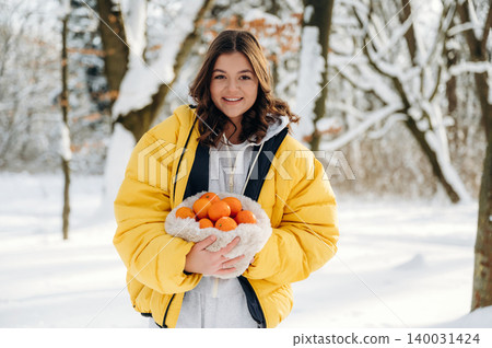 Young woman with brown hair wearing a yellow puffer jacket is holding a bowl of oranges in a snowy forest setting with trees and snow-covered ground surrounding her Young woman with brown hair wearing a yellow puffer jacket is holding a bowl of oranges in a snowy forest setting with trees and snow-covered ground surrounding her 140031424