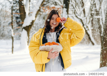 Young woman in a bright yellow puffer jacket holds a basket of oranges while standing in a snowy forest, snowflakes falling around her in a winter scene Young woman in a bright yellow puffer jacket holds a basket of oranges while standing in a snowy forest, snowflakes falling around her in a winter scene 140031442