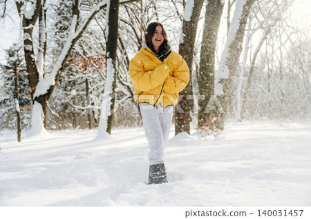 Young woman wearing a bright yellow puffer jacket and gray pants is walking through a snowy forest, surrounded by tall trees and falling snowflakes during winter Young woman wearing a bright yellow puffer jacket and gray pants is walking through a snowy forest, surrounded by tall trees and falling snowflakes during winter 140031457