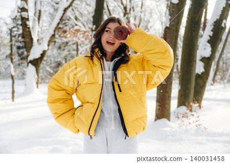 Young woman wearing a bright yellow puffer jacket and gray sweatpants is playfully holding a red ornament while standing in a snowy forest during winter Young woman wearing a bright yellow puffer jacket and gray sweatpants is playfully holding a red ornament while standing in a snowy forest during winter 140031458