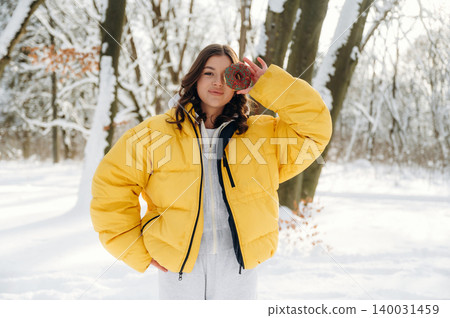 Young woman wearing a bright yellow puffer jacket stands in a snowy forest holding a red ornament, surrounded by snow-covered trees and a winter landscape with soft sunlight Young woman wearing a bright yellow puffer jacket stands in a snowy forest holding a red ornament, surrounded by snow-covered trees and a winter landscape with soft sunlight 140031459
