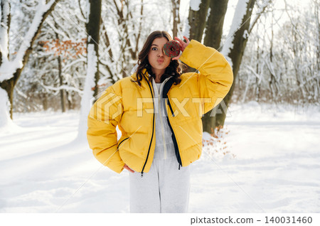 Young woman in a bright yellow puffer jacket poses playfully in a snowy forest, holding a cup while surrounded by snow-covered trees and a winter landscape with soft sunlight Young woman in a bright yellow puffer jacket poses playfully in a snowy forest, holding a cup while surrounded by snow-covered trees and a winter landscape with soft sunlight 140031460