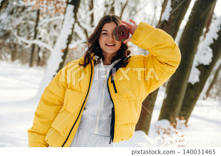 Young woman with brown hair wearing a bright yellow puffer jacket is playfully holding a red ornament in a snowy forest setting during winter Young woman with brown hair wearing a bright yellow puffer jacket is playfully holding a red ornament in a snowy forest setting during winter 140031465