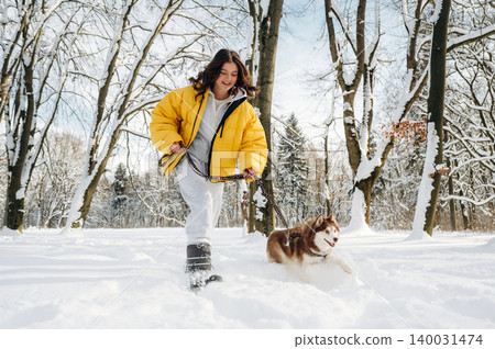 Young woman in a yellow puffer jacket is walking a brown and white dog through a snowy park with trees in the background during wintertime outdoor activity Young woman in a yellow puffer jacket is walking a brown and white dog through a snowy park with trees in the background during wintertime outdoor activity 140031474