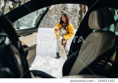 Woman in yellow winter jacket stands outside a car holding a Siberian Husky dog in a snowy landscape with trees in the background during a winter day 140031481