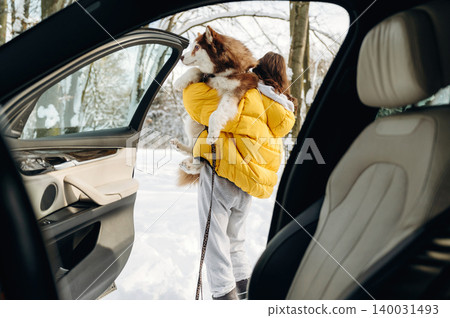Woman in yellow jacket carrying a dog steps out of a car into a snowy landscape surrounded by trees, showcasing a winter outdoor adventure with copy space Woman in yellow jacket carrying a dog steps out of a car into a snowy landscape surrounded by trees, showcasing a winter outdoor adventure with copy space 140031493