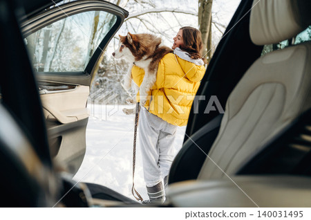 Woman in yellow puffer jacket carrying a dog stands outside a car in a snowy landscape with trees, showcasing a winter outing and pet companionship Woman in yellow puffer jacket carrying a dog stands outside a car in a snowy landscape with trees, showcasing a winter outing and pet companionship 140031495