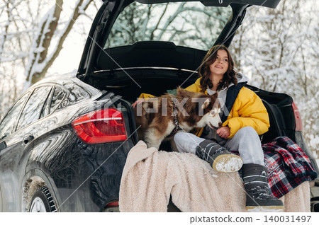 Young woman in yellow winter jacket sits in car trunk with her dog, surrounded by snow-covered trees, enjoying a cozy moment during a winter outing in nature 140031497