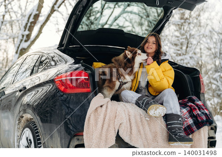 Young woman in yellow winter jacket sits in the trunk of a black SUV with a brown dog during a snowy day in a forested area, enjoying the winter scenery and companionship Young woman in yellow winter jacket sits in the trunk of a black SUV with a brown dog during a snowy day in a forested area, enjoying the winter scenery and companionship 140031498