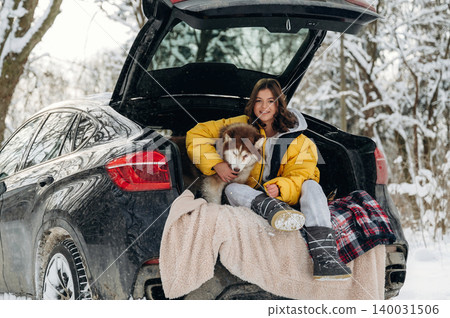 Young girl with brown hair wearing a yellow puffer jacket is sitting in the trunk of a black SUV with a dog in a snowy landscape during winter Young girl with brown hair wearing a yellow puffer jacket is sitting in the trunk of a black SUV with a dog in a snowy landscape during winter 140031506