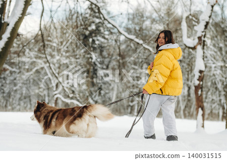 Young woman wearing a bright yellow puffer jacket walks her fluffy dog on a snowy path surrounded by trees, showcasing a winter outdoor scene with a playful atmosphere Young woman wearing a bright yellow puffer jacket walks her fluffy dog on a snowy path surrounded by trees, showcasing a winter outdoor scene with a playful atmosphere 140031515