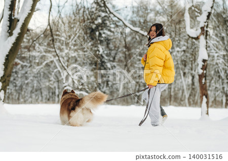 Young woman in a bright yellow puffer jacket walks a fluffy dog on a leash through a snowy landscape with trees and a winter sky in the background Young woman in a bright yellow puffer jacket walks a fluffy dog on a leash through a snowy landscape with trees and a winter sky in the background 140031516