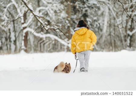 Child wearing a bright yellow puffer jacket walks a small dog on a leash through a snowy landscape with trees in the background during winter season Child wearing a bright yellow puffer jacket walks a small dog on a leash through a snowy landscape with trees in the background during winter season 140031522
