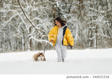 Young woman in a yellow puffer jacket walks a dog on a leash through a snowy landscape with trees in the background during winter outdoor activity Young woman in a yellow puffer jacket walks a dog on a leash through a snowy landscape with trees in the background during winter outdoor activity 140031526