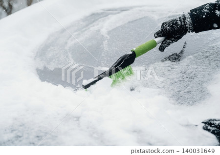 Hand wearing black glove using a green snow brush to clear snow from a car windshield covered in white snow during winter weather conditions in an outdoor setting Hand wearing black glove using a green snow brush to clear snow from a car windshield covered in white snow during winter weather conditions in an outdoor setting 140031649