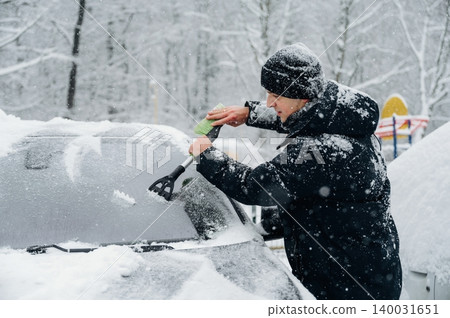 Man in black winter coat is scraping snow off a car windshield with a green ice scraper in a snowy parking lot surrounded by trees during winter weather Man in black winter coat is scraping snow off a car windshield with a green ice scraper in a snowy parking lot surrounded by trees during winter weather 140031651