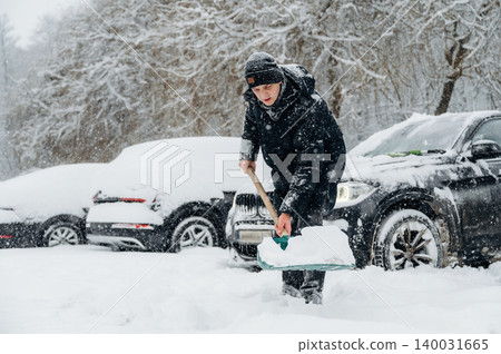 Young male shoveling snow in a winter landscape, wearing a black coat and hat, with parked cars covered in snow and trees in the background during a snowy day Young male shoveling snow in a winter landscape, wearing a black coat and hat, with parked cars covered in snow and trees in the background during a snowy day 140031665