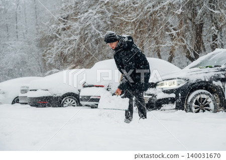 Man in black coat and hat shoveling snow in a parking lot covered with snow, several cars partially buried in the background during winter weather conditions Man in black coat and hat shoveling snow in a parking lot covered with snow, several cars partially buried in the background during winter weather conditions 140031670