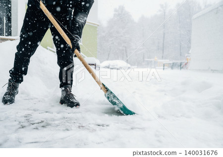 Individual wearing black pants and boots is shoveling snow with a green shovel during a heavy snowfall in a residential area with trees in the background and a building nearby Individual wearing black pants and boots is shoveling snow with a green shovel during a heavy snowfall in a residential area with trees in the background and a building nearby 140031676