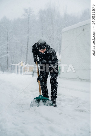Individual dressed in black winter clothing is shoveling snow from a pathway during a heavy snowfall, surrounded by trees and a white building in a winter landscape 140031679