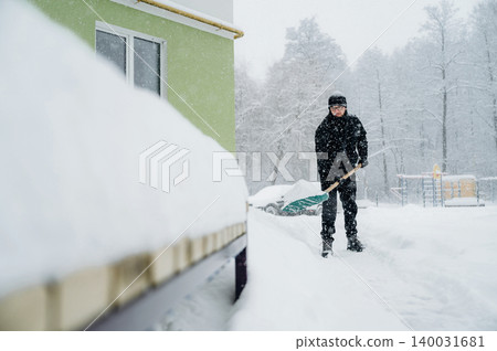 Man dressed in black winter clothing shoveling snow from a sidewalk near a green building during a heavy snowfall with trees and playground visible in the background Man dressed in black winter clothing shoveling snow from a sidewalk near a green building during a heavy snowfall with trees and playground visible in the background 140031681