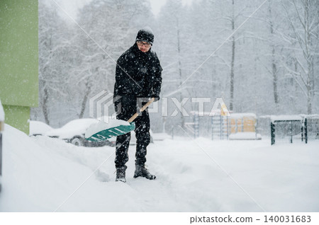 Man in black winter clothing shoveling snow with a green shovel during a heavy snowfall near a playground and parked cars in a residential area Man in black winter clothing shoveling snow with a green shovel during a heavy snowfall near a playground and parked cars in a residential area 140031683