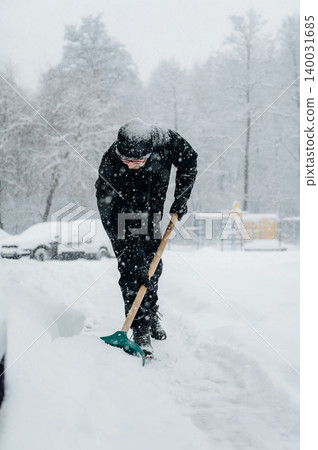 Individual dressed in black winter clothing is shoveling snow from a pathway during a heavy snowfall in a residential area with trees and parked cars in the background Individual dressed in black winter clothing is shoveling snow from a pathway during a heavy snowfall in a residential area with trees and parked cars in the background 140031685
