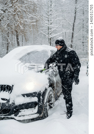 Man dressed in black winter clothing is clearing snow from a car parked in a snowy landscape with trees covered in snow during a winter storm Man dressed in black winter clothing is clearing snow from a car parked in a snowy landscape with trees covered in snow during a winter storm 140031705