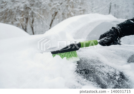 Person using a snow brush to clear snow from a car roof during a winter snowfall, with trees and a snowy landscape visible in the background 140031715