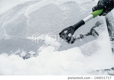 Person using a black and green snow scraper to remove snow from a car windshield covered in white snow during winter weather conditions in a cold environment Person using a black and green snow scraper to remove snow from a car windshield covered in white snow during winter weather conditions in a cold environment 140031717