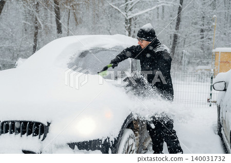 Man in black winter clothing is clearing snow from a car windshield during a snowfall in a wooded area with trees and a fence visible in the background Man in black winter clothing is clearing snow from a car windshield during a snowfall in a wooded area with trees and a fence visible in the background 140031732