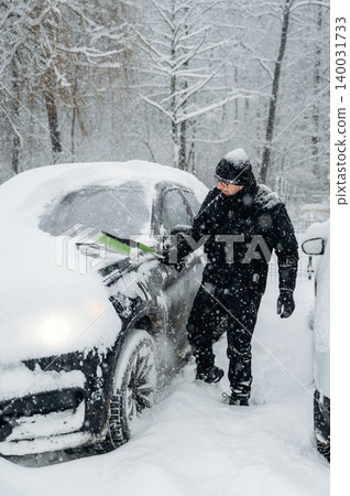 Man in black winter clothing is clearing snow from a car parked in a snowy landscape with trees in the background during a heavy snowfall Man in black winter clothing is clearing snow from a car parked in a snowy landscape with trees in the background during a heavy snowfall 140031733