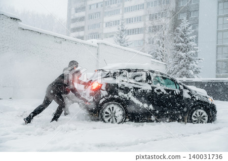 Individual pushing a black car stuck in deep snow during a heavy winter snowstorm, surrounded by snow-covered trees and a tall building in the background with limited visibility Individual pushing a black car stuck in deep snow during a heavy winter snowstorm, surrounded by snow-covered trees and a tall building in the background with limited visibility 140031736