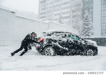 Individual dressed in black winter clothing is pushing a snow-covered car stuck in deep snow during a heavy snowfall in an urban environment with tall buildings visible Individual dressed in black winter clothing is pushing a snow-covered car stuck in deep snow during a heavy snowfall in an urban environment with tall buildings visible 140031737