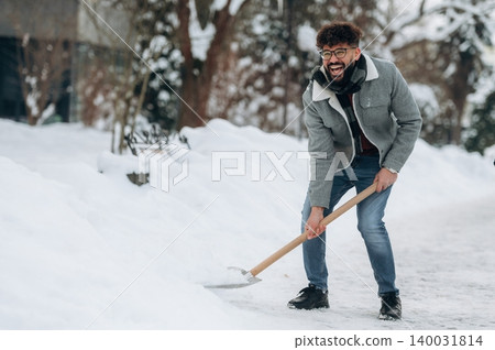 Young man with curly hair wearing a gray jacket and scarf is shoveling snow on a winter day, surrounded by snow-covered ground and trees in the background Young man with curly hair wearing a gray jacket and scarf is shoveling snow on a winter day, surrounded by snow-covered ground and trees in the background 140031814