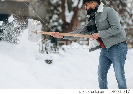 Man with beard shoveling snow in winter attire using a metal shovel, surrounded by a snowy landscape and trees in the background during a cold day Man with beard shoveling snow in winter attire using a metal shovel, surrounded by a snowy landscape and trees in the background during a cold day 140031817