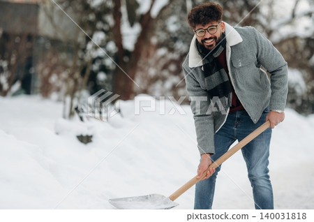 Young man with curly hair wearing glasses and a gray jacket is shoveling snow in a winter landscape with trees and a pathway visible in the background 140031818