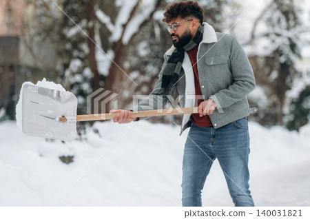 Man with curly hair wearing a gray jacket and scarf is shoveling snow with a metal shovel in a winter landscape surrounded by trees and snow-covered ground Man with curly hair wearing a gray jacket and scarf is shoveling snow with a metal shovel in a winter landscape surrounded by trees and snow-covered ground 140031821