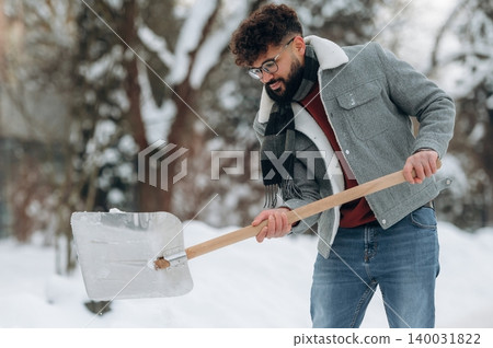 Man with curly hair and glasses shoveling snow in a winter landscape wearing a gray jacket and scarf, surrounded by trees and a snowy ground during a cold day Man with curly hair and glasses shoveling snow in a winter landscape wearing a gray jacket and scarf, surrounded by trees and a snowy ground during a cold day 140031822
