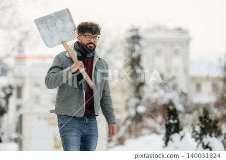 Man with curly hair wearing glasses and a gray jacket is walking outdoors in the snow while holding a shovel, trees and buildings visible in the background during winter 140031824