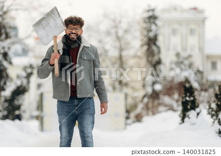 Smiling man with curly hair wearing a gray jacket and scarf is walking through a snowy landscape holding a shovel, trees and buildings visible in the background 140031825
