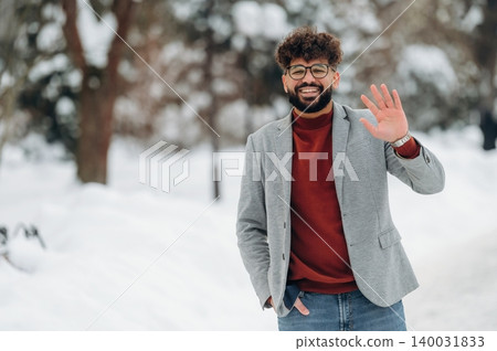 Young man with curly hair wearing a gray blazer and red sweater is smiling and waving in a snowy outdoor park setting with trees in the background during winter Young man with curly hair wearing a gray blazer and red sweater is smiling and waving in a snowy outdoor park setting with trees in the background during winter 140031833