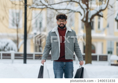 Man with curly hair wearing a gray jacket and glasses is holding shopping bags while standing in a snowy park with trees and buildings in the background during winter Man with curly hair wearing a gray jacket and glasses is holding shopping bags while standing in a snowy park with trees and buildings in the background during winter 140031838