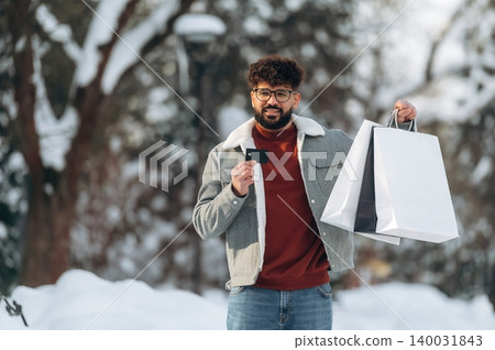 Young man with curly hair wearing glasses is standing outdoors in winter, holding shopping bags and a credit card in a snowy park with trees in the background 140031843