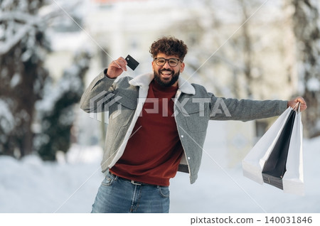 Young man with curly hair wearing a gray jacket and red sweater is joyfully holding shopping bags and a credit card while standing in a snowy outdoor setting with trees in the background Young man with curly hair wearing a gray jacket and red sweater is joyfully holding shopping bags and a credit card while standing in a snowy outdoor setting with trees in the background 140031846